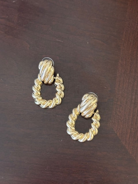A pair of gold-toned twisted rope hoop earrings with a textured, ribbed center, displayed on a dark brown wooden surface.